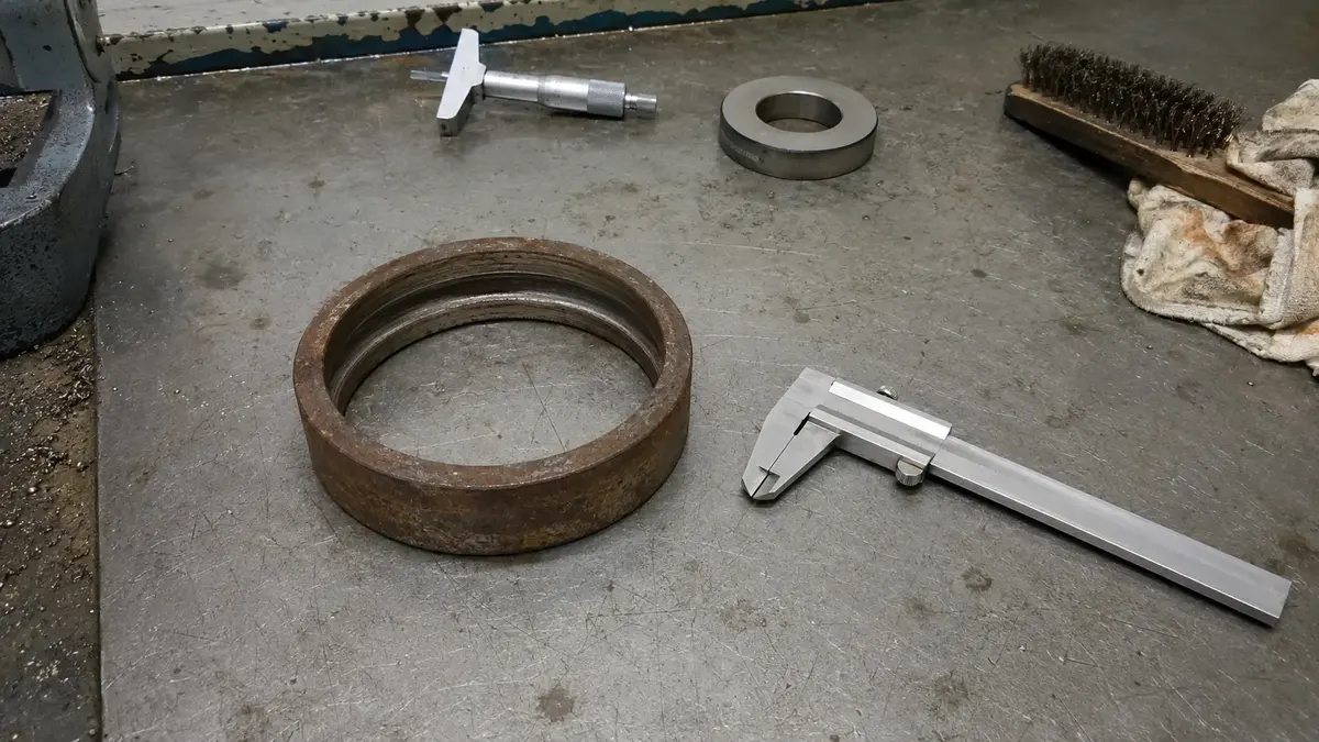 A corroded deep groove ball bearing outer ring on a QC bench with its stamping band worn smooth and blank while the ball groove in the bore is still intact, calipers and a depth micrometer lying beside it: measurement recovers the basic number but not the seal, cage, clearance or tolerance class