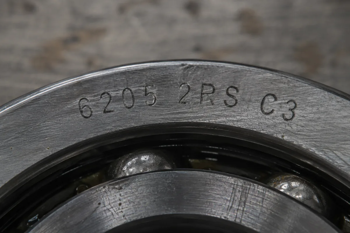 Macro close-up of a deep groove ball bearing outer ring laser-etched with the full designation 6205 2RS C3, with the rolling elements visible below the marking