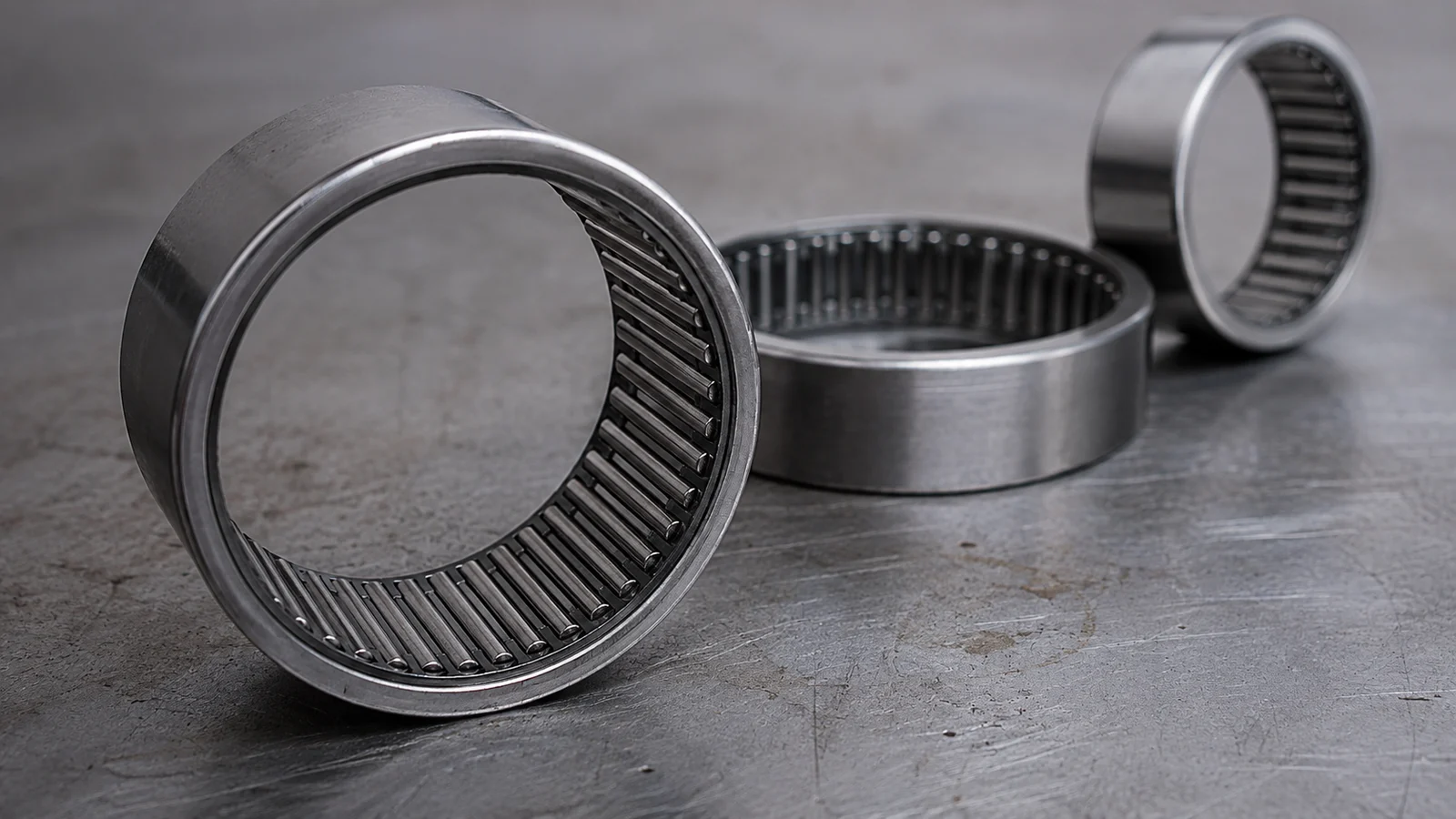 Three machined-ring needle roller bearings on a steel workbench, the nearest tipped on its side to reveal the dense ring of slender needle rollers inside its bore