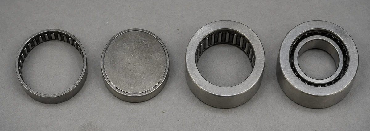 Four needle roller bearings photographed from above in a row: an open-end drawn cup and a closed-end drawn cup with thin stamped walls, then a machined-ring bearing and a machined-ring bearing with an inner ring, both with visibly thicker walls